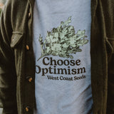 A person wearing choose optimism sweatshirt west coast seeds beneath an overshirt